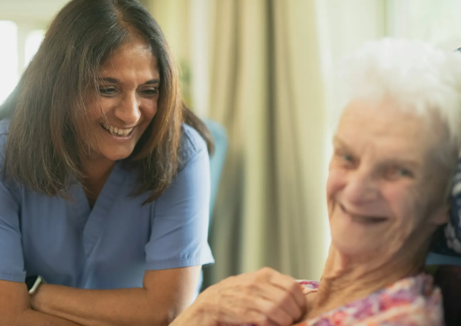 A carer in a blue tunic laughing with an older woman in her own front room