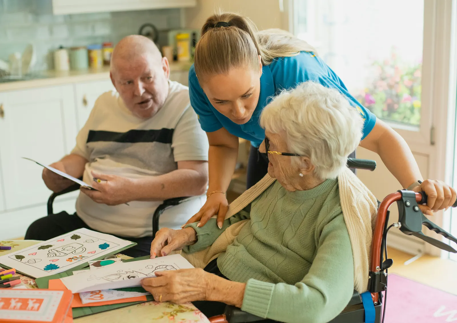 A carer leaning in to help an older woman at a kitchen table, working through something together